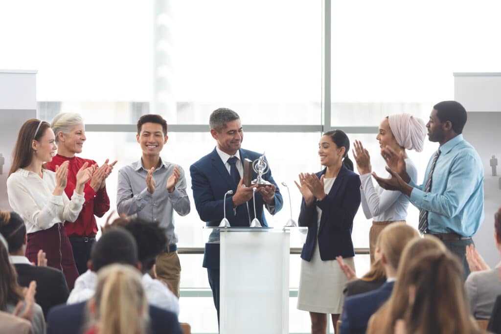 businessman holding award on podium with colleagues in front of business professionals at business seminar in office building for employee engagement